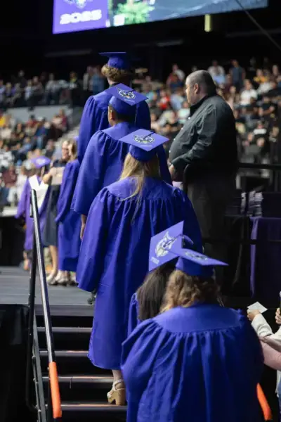 Sinistes rõivastes lõpetajad tõusevad tseremoonial lavale, seljas kaunistatud mütsid. Taustal vaatab rahvahulk, mis loob piduliku õhkkonna.